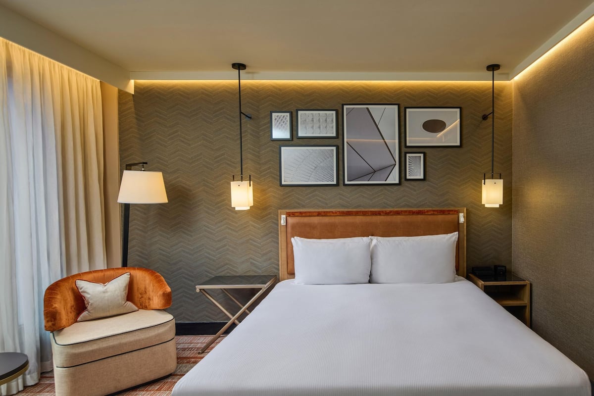 Habitación de hotel con cama, una silla, una lámpara y cuadros en la pared.