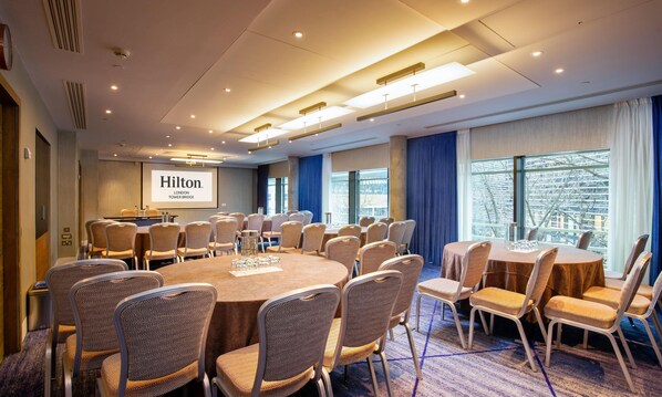 A conference room with round tables and chairs, a projector screen displaying 'Hilton London Tower Bridge', and large windows with a view of the city. - Hilton London Tower Bridge (London)