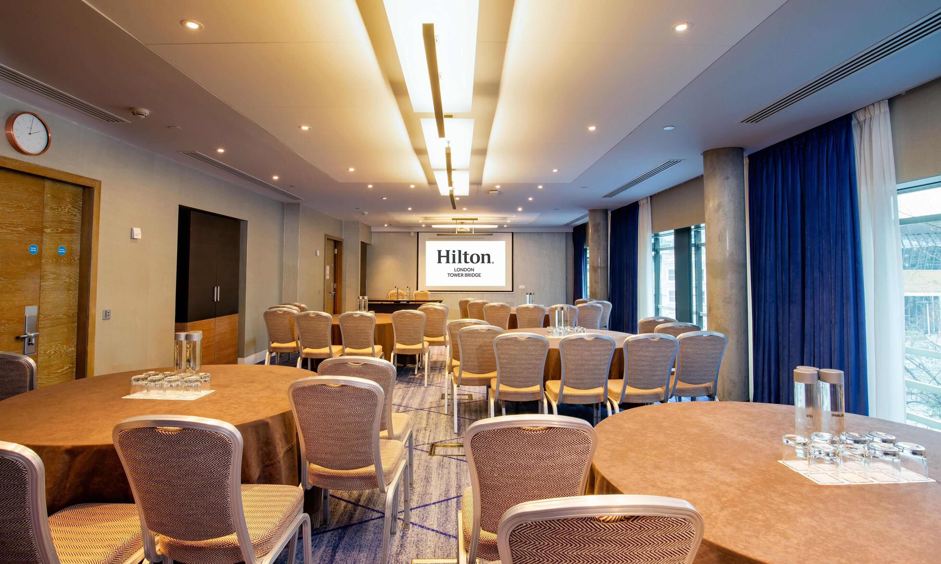 A conference room with a large screen displaying 'Hilton London Tower Bridge,' multiple round tables with chairs, and a clock on the left wall.