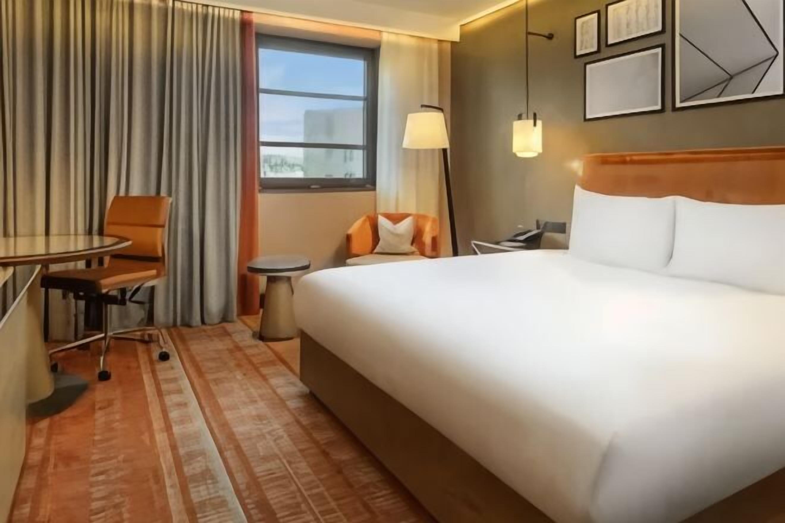 A hotel room with a large bed, a desk, a chair, a small table, a lamp, and a window.