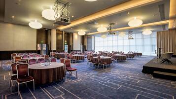 A large conference room with round tables and chairs, a stage, and a high ceiling with modern lighting fixtures.