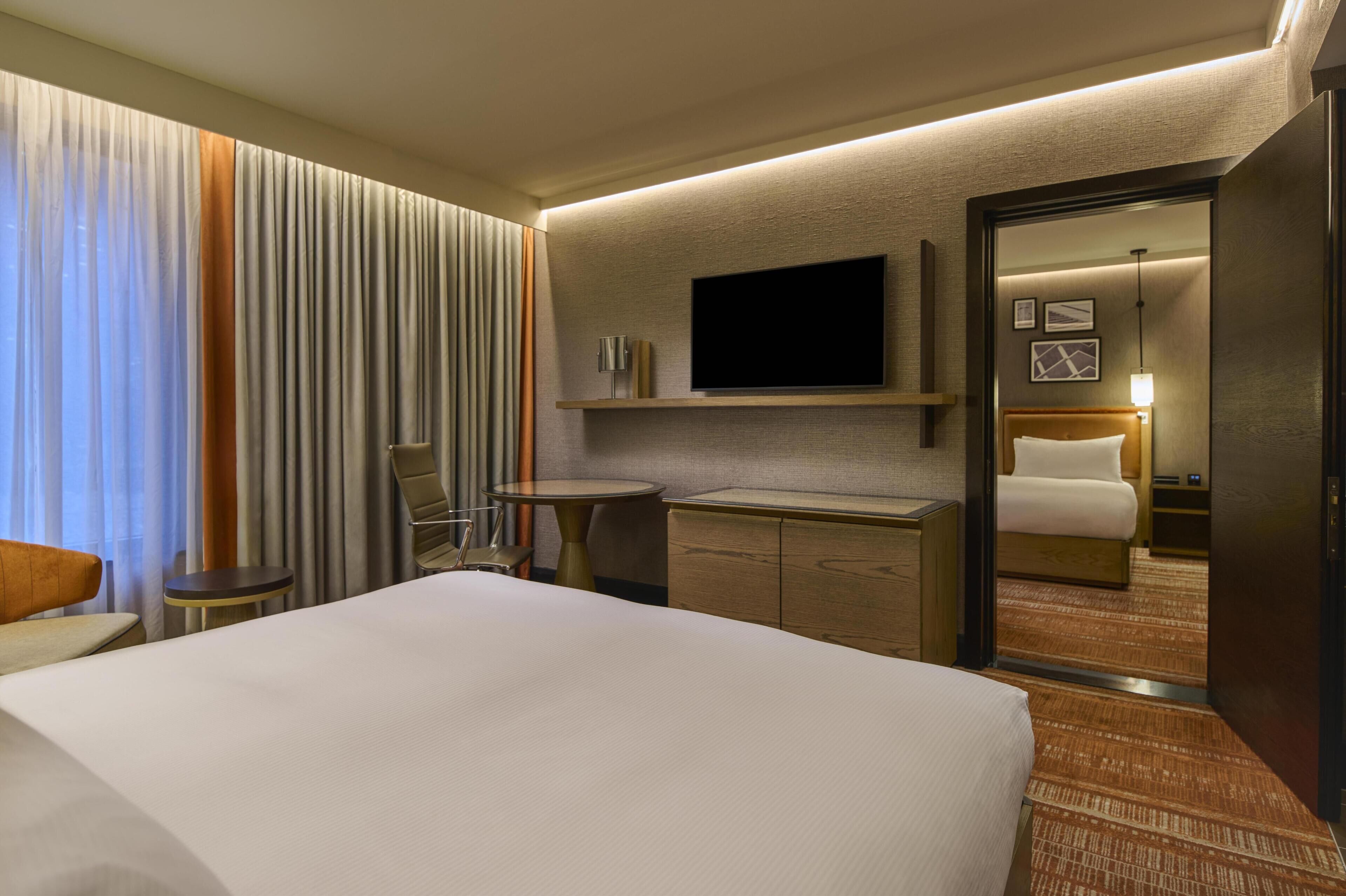 A hotel room with a large bed, a television mounted on the wall, a small round table, a cabinet, and a mirror reflecting the room.