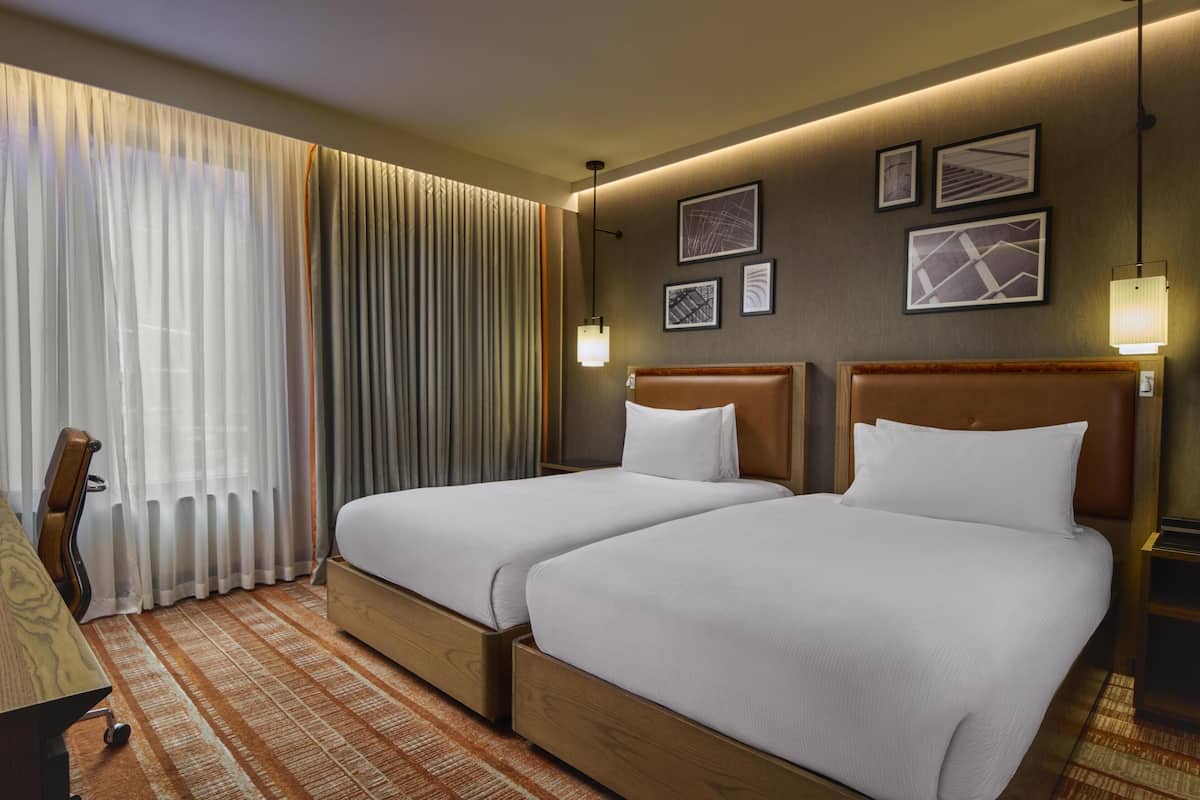 Habitación de hotel con dos camas, un escritorio, una silla y un ventanal grande con cortinas.