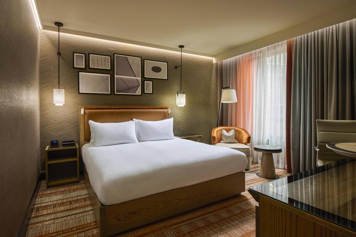 Habitación de hotel con una cama grande, mesitas de noche, un escritorio con silla, un espejo y cuadros en la pared.