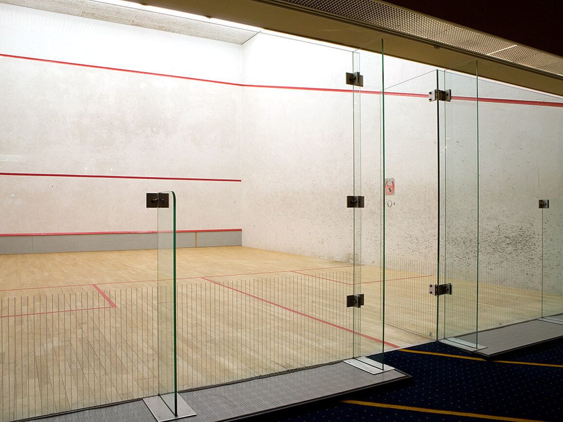 Un court de squash avec des murs en verre, un plancher en bois et une zone recouverte d’une moquette bleue.