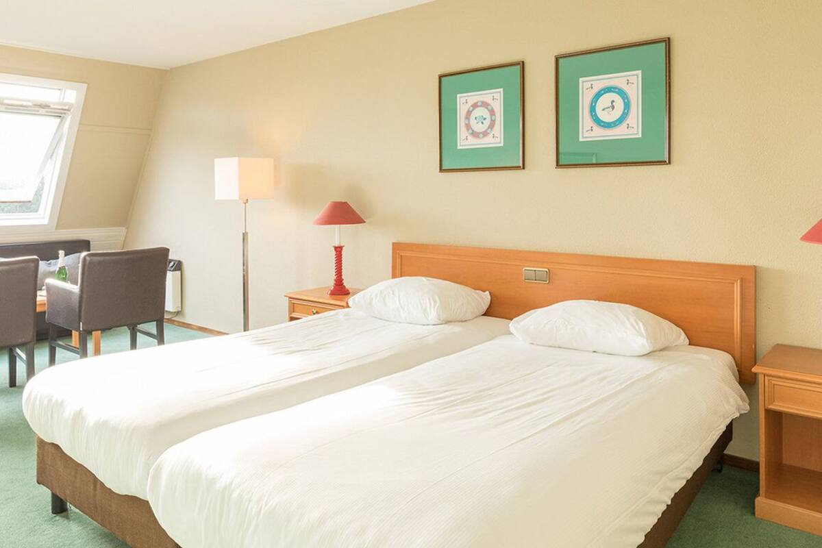 Ein Hotelzimmer mit zwei Betten, einem Schreibtisch mit Stühlen, einer Lampe und zwei gerahmten Uhren an der Wand.