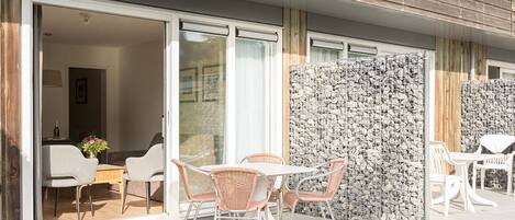 Un patio al aire libre con una mesa, sillas, un muro de madera y un ventanal corredera que da a un espacio interior.