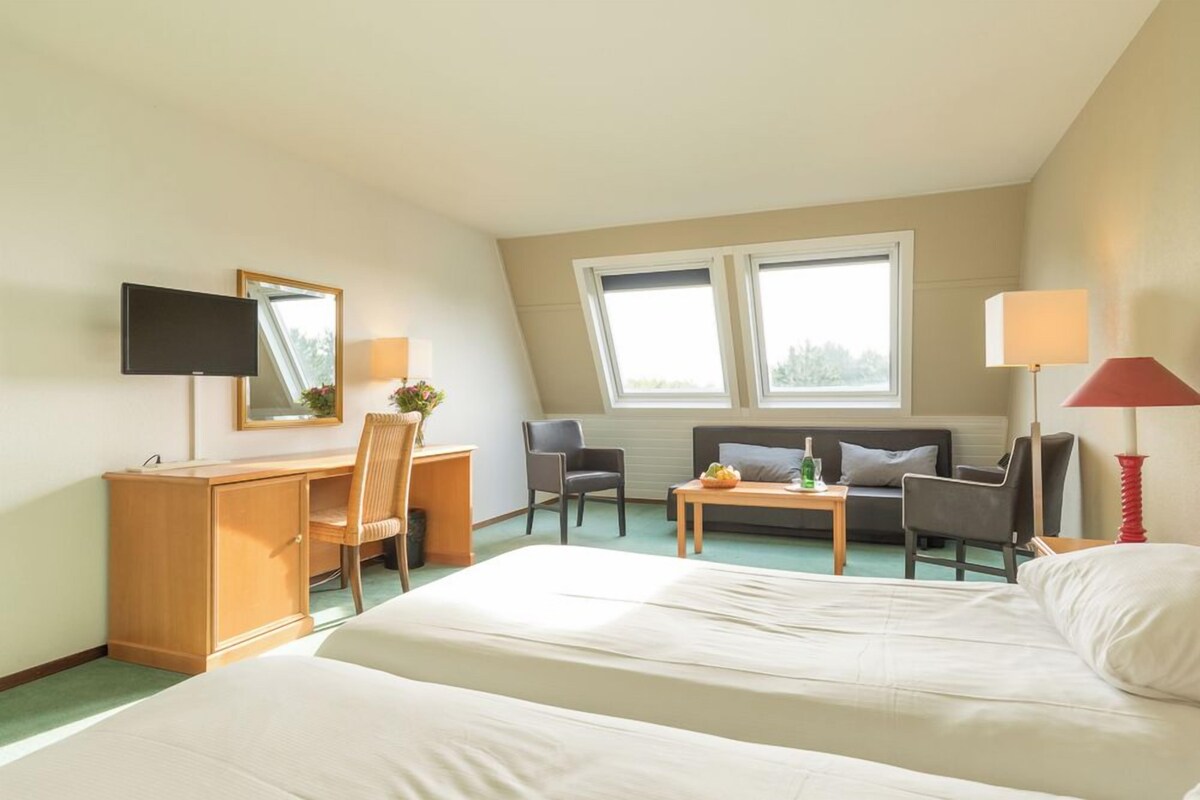 Quarto de hotel com cama, escrivaninha, cadeira, sofá e duas janelas.