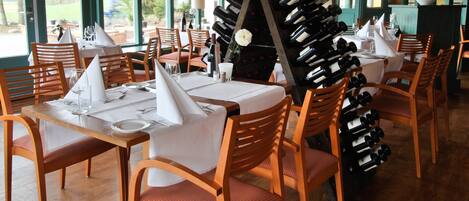 A restaurant with wooden chairs and tables, a wine rack, and large windows with a view of the outdoors.