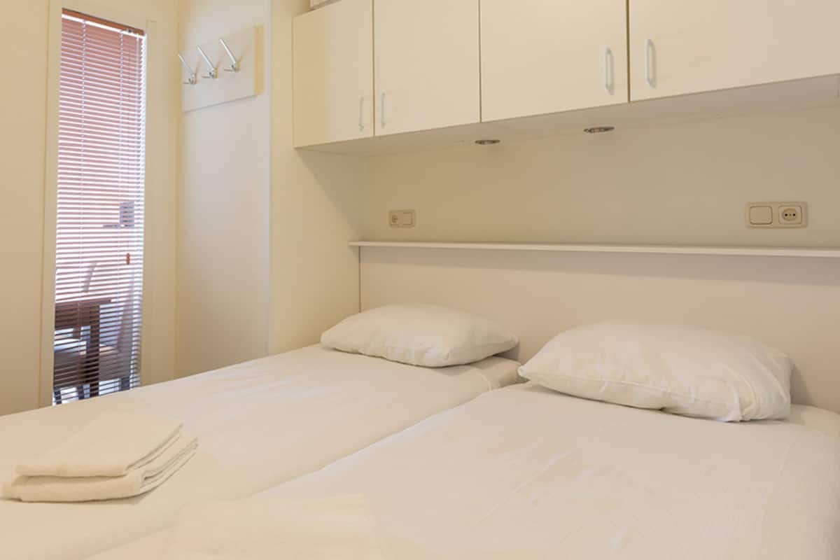 Ein Schlafzimmer mit einem Bett, zwei Kissen, einem gefalteten Handtuch, einem Fenster mit Jalousien und einem an der Wand befestigten Regal.