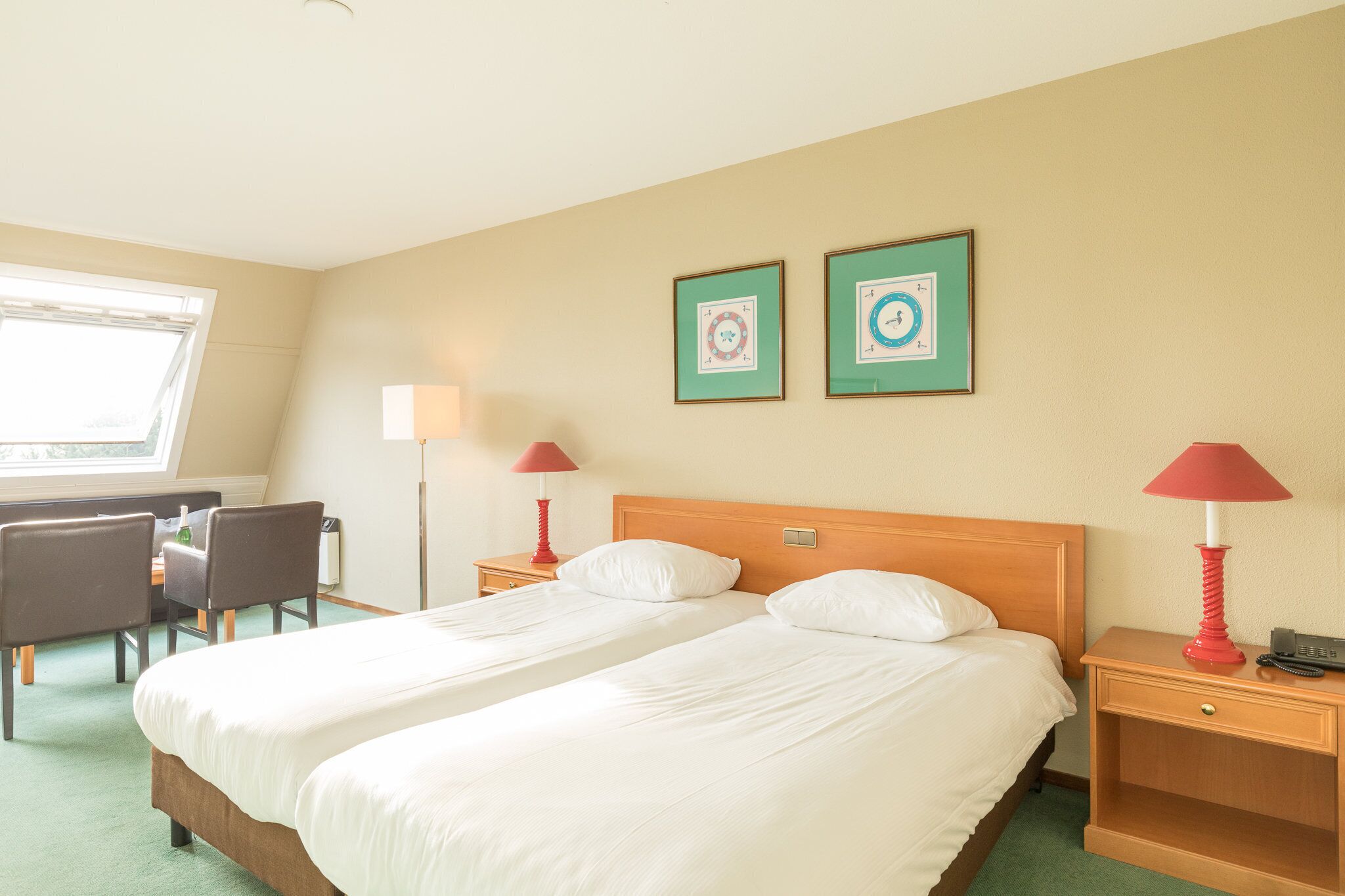 A hotel room with two single beds, a desk with chairs, a lamp, and two framed pictures on the wall.