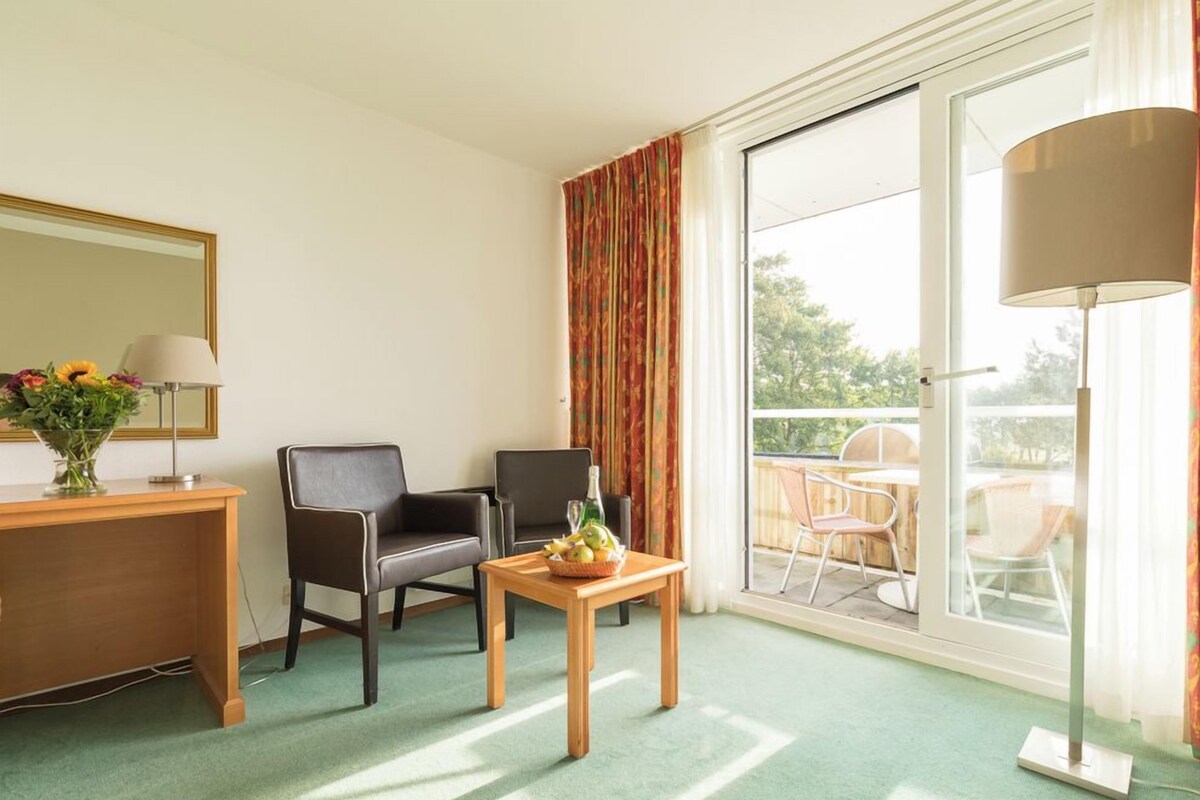 A hotel room with a desk, two chairs, a small table with fruit, a lamp, and a large window with a view.