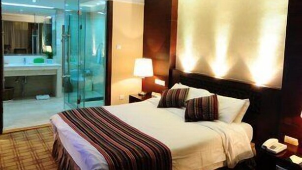 A hotel room with a bed, bedside tables, a nightstand, a lamp, and a view of a bathroom with a glass shower enclosure. - Yunnan Aviation Sightseeing Hotel (Lijiang)