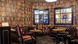 Um salão estilo biblioteca, com estantes de livros, mobiliário de couro e duas janelas.