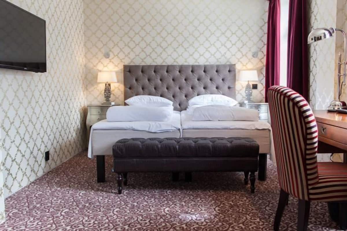 Un dormitorio con una cama con cabecera capitonada, una mesita de noche con una lámpara, una silla a rayas y un aparador de madera.