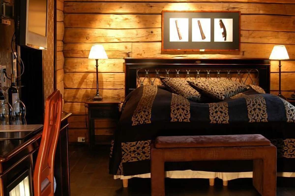 Una habitación tipo cabaña de madera con una cama, dos lámparas de noche, un cuadro enmarcado sobre la cama y un banco de madera.