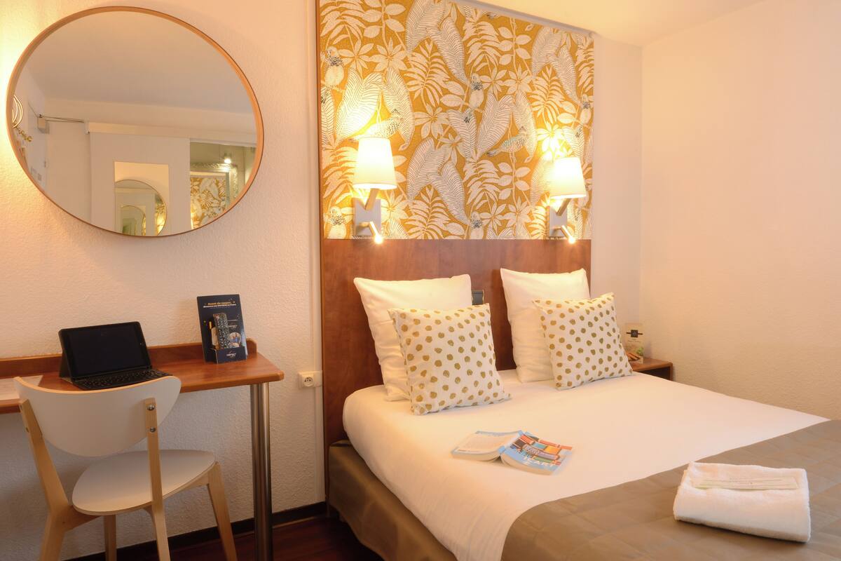 A hotel room with a bed, a desk with a laptop, a chair, a round mirror, and a patterned headboard.