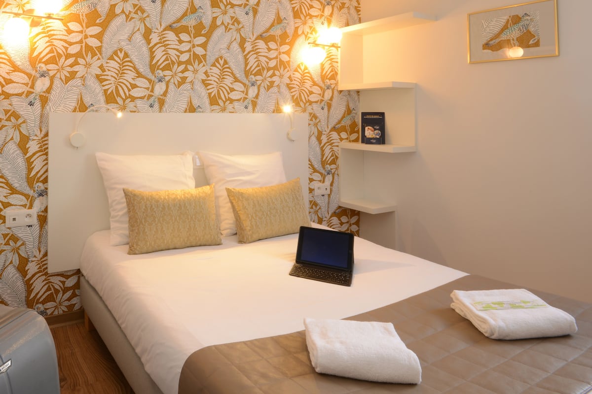 A hotel room with a bed, a laptop, a bedside table, and a wall with a leaf-patterned wallpaper.