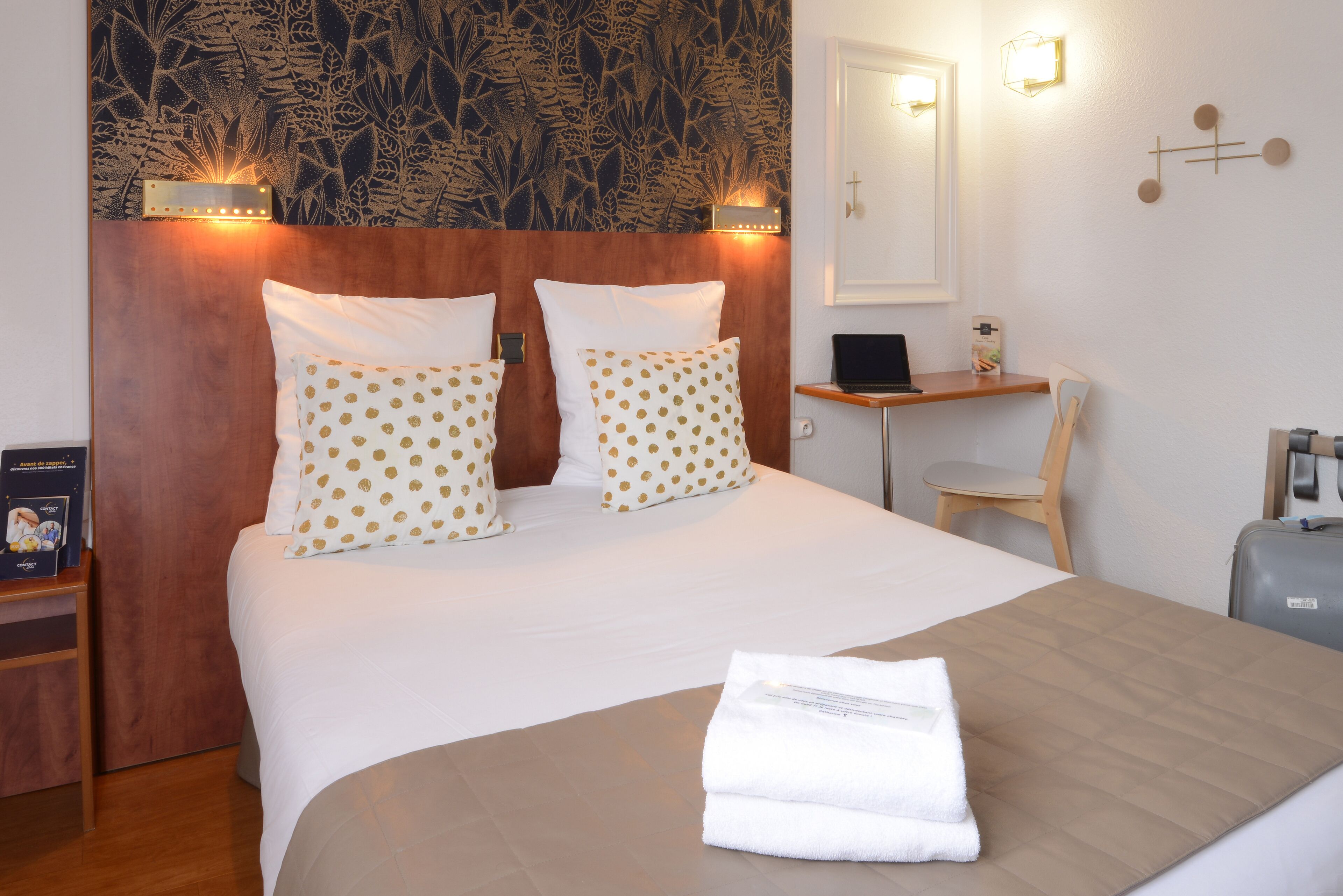 A neatly made bed with white and gold polka dot pillows, a wooden headboard, a bedside table with a laptop, and a small desk with a chair.