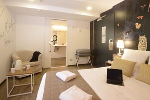 Habitación de hotel con cama, laptop, silla, una mesita con tetera y tazas, y baño visible a través de una puerta abierta.