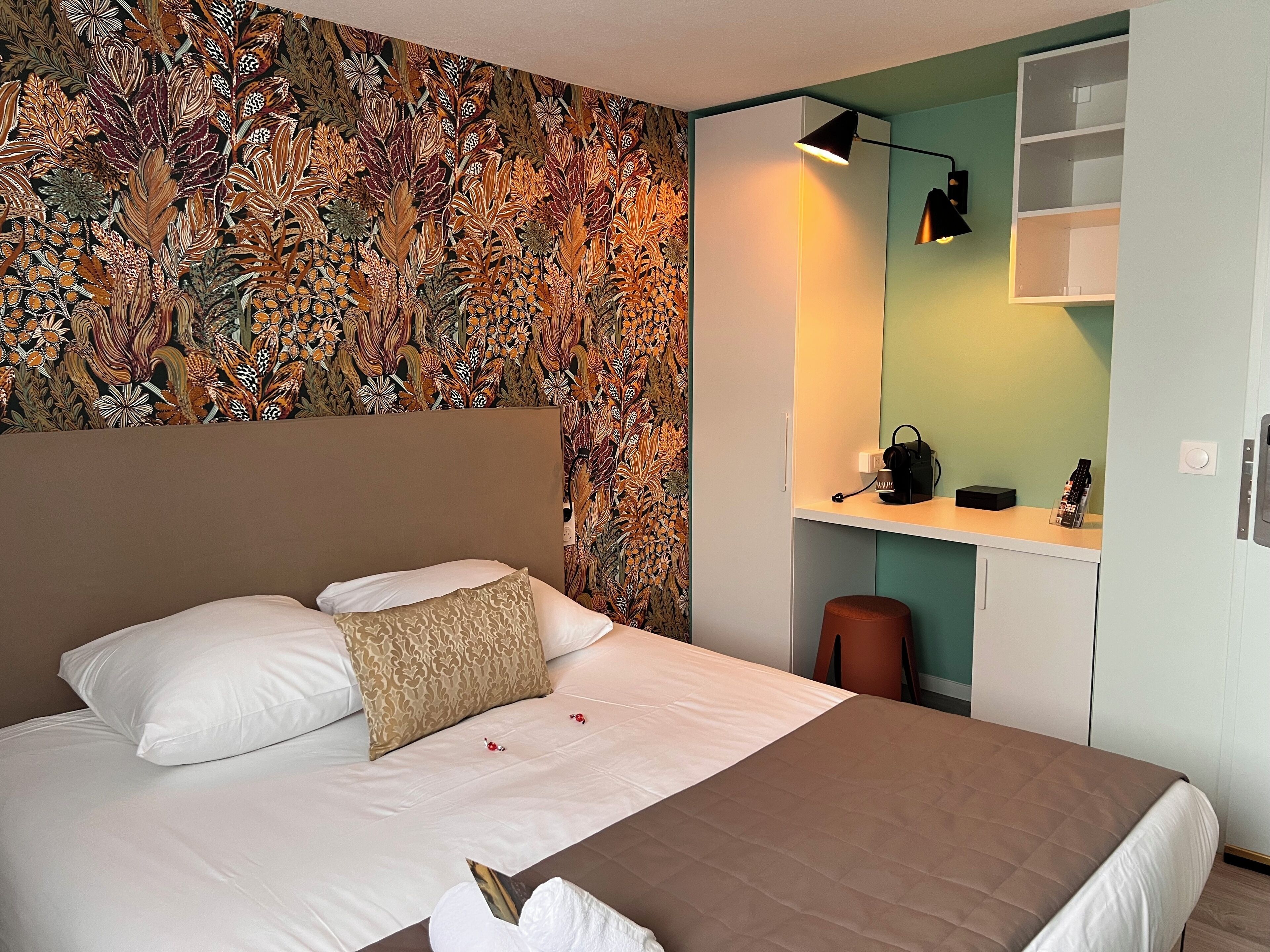 A bedroom with a large bed, a headboard, a patterned wallpaper, a desk with a lamp, and a small stool.