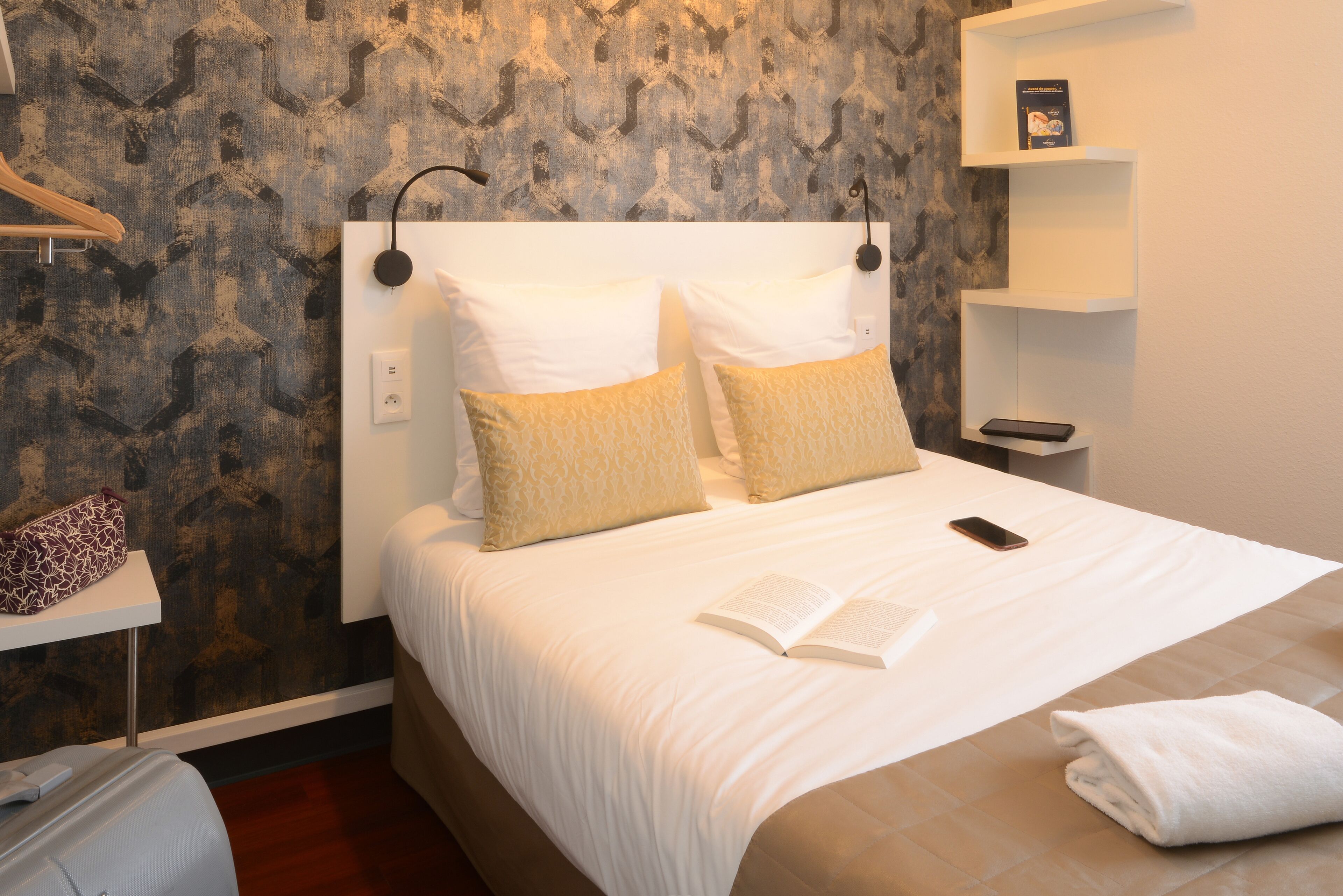 A neatly made bed with a white duvet, two pillows, and a book on it. There is a white bedside table with a phone and a towel on it. The wall has a textured pattern.