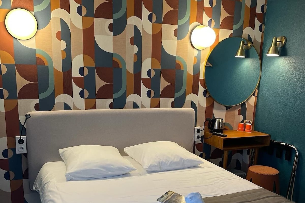 A neatly made bed with white linens, a grey headboard, and a blue blanket. There is a small wooden nightstand with a kettle and a cup. The wall features a colorful abstract pattern.