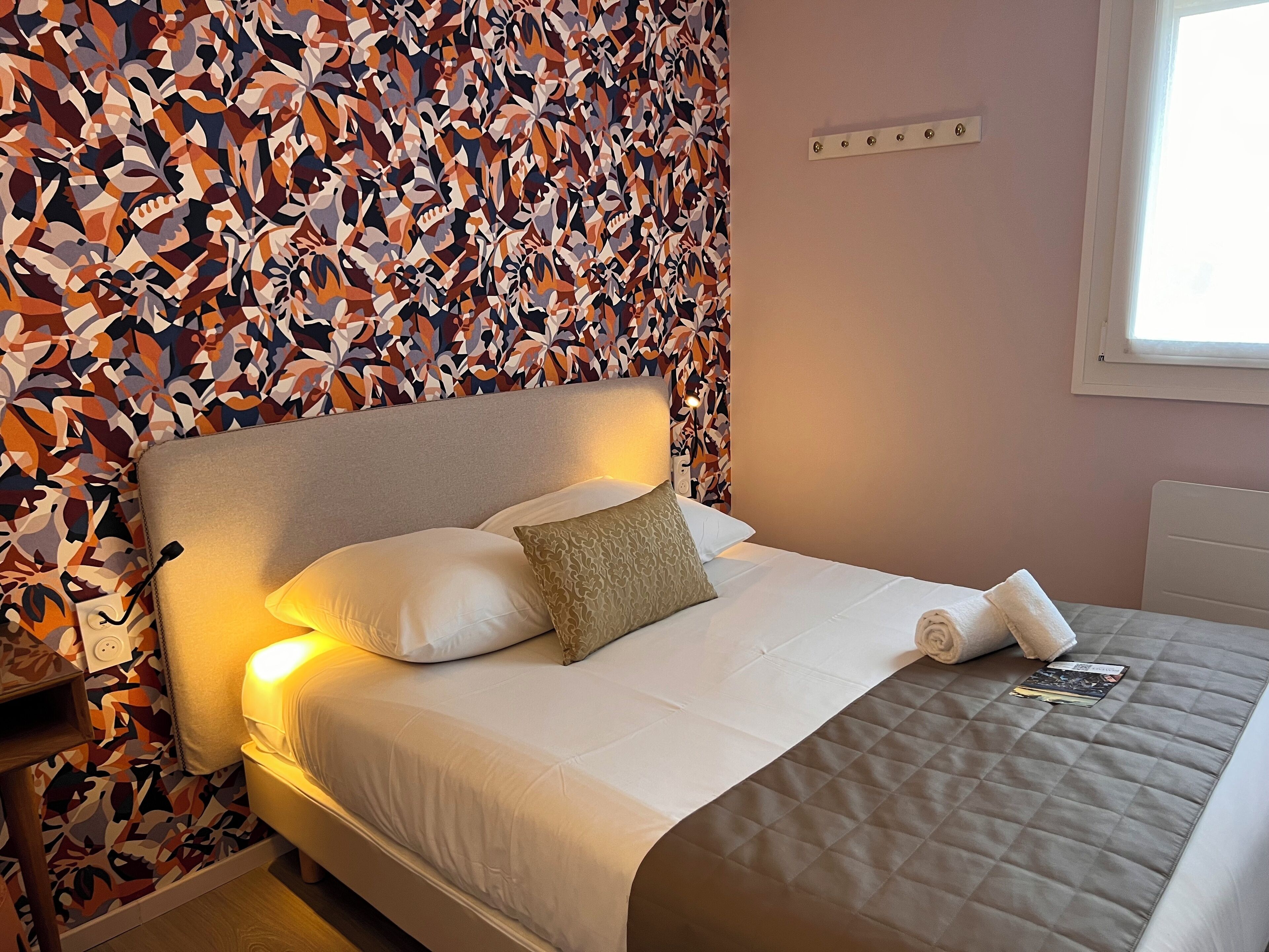 Una cama bien hecha con sábanas blancas y un cubrecama acolchado, un papel tapiz estampado en el fondo y una ventana a la derecha.