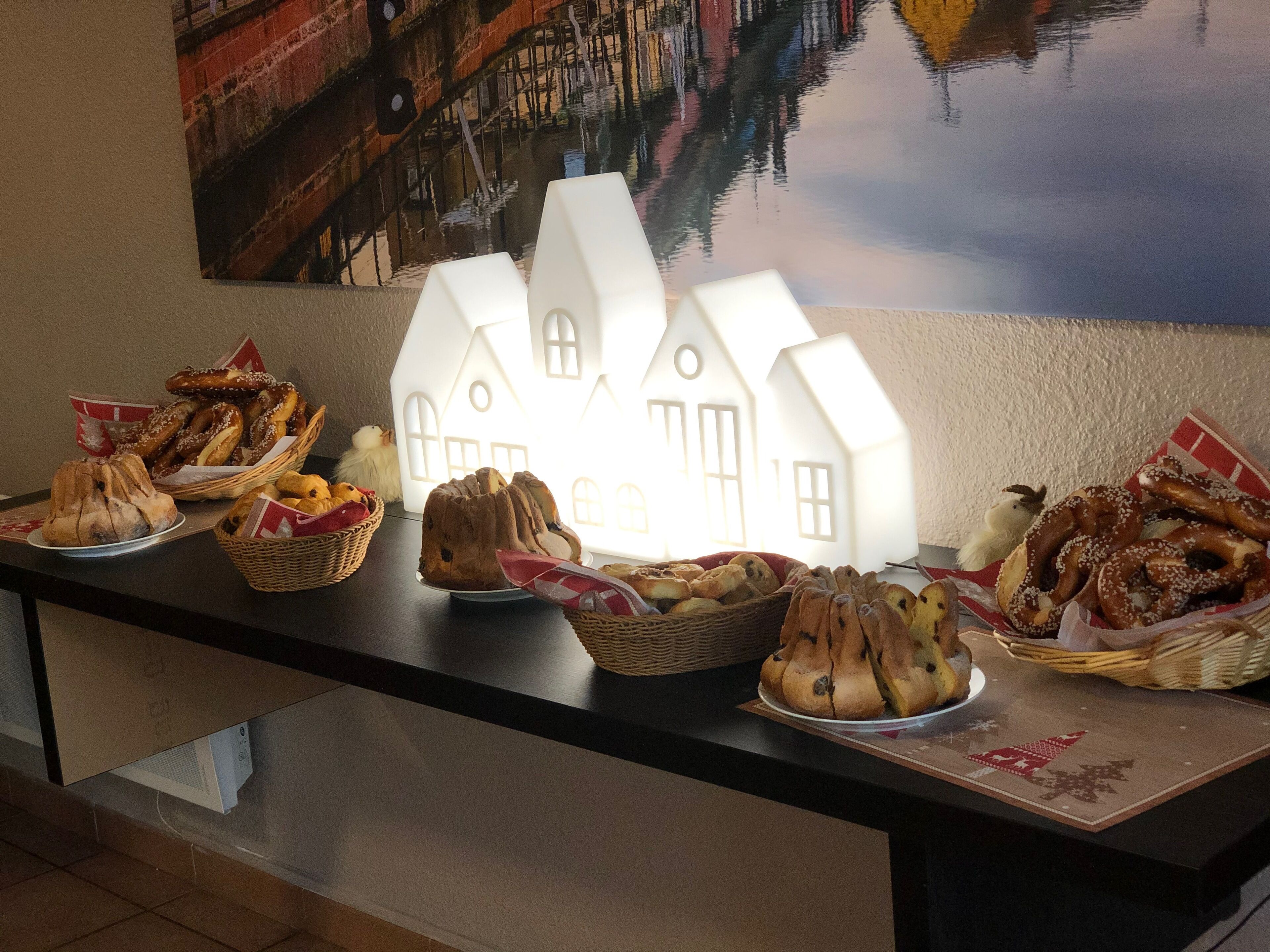 A buffet table with various breads and pastries, including pretzels and a loaf of bread, set against a backdrop of a wall with a painted scene and a white house-shaped light fixture.