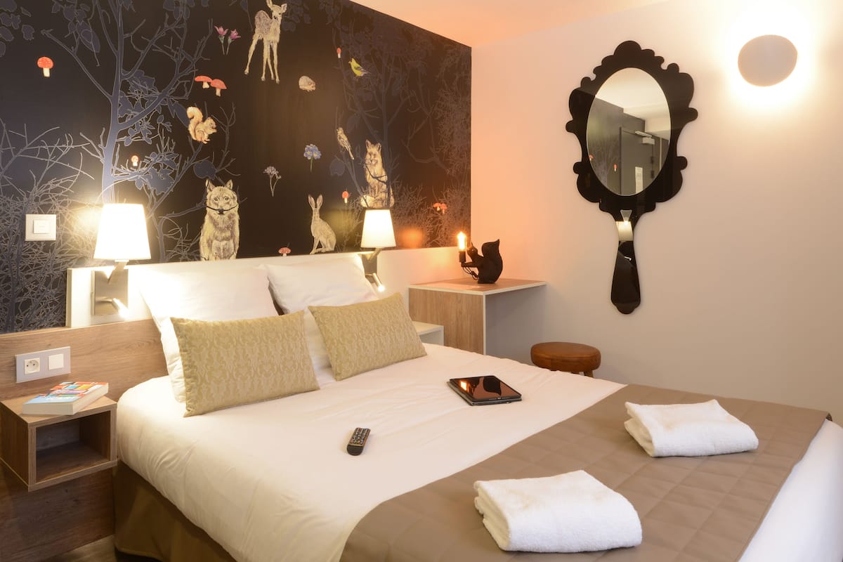 A neatly made bed with pillows, a bedside table with a lamp, a mirror, and a wall with a nature-themed mural.
