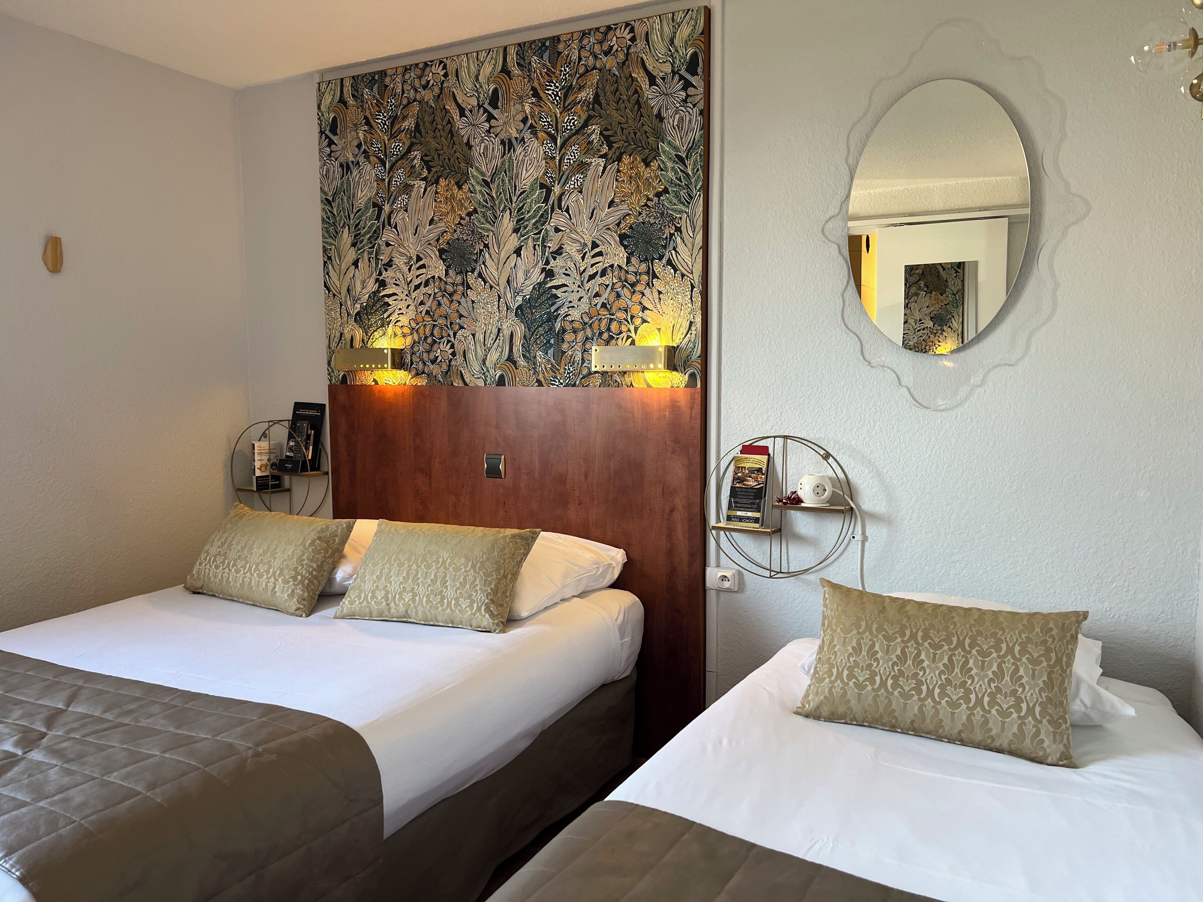 A hotel room with two beds, a floral-patterned headboard, bedside lamps, a round mirror, and a small shelf with a coffee maker.