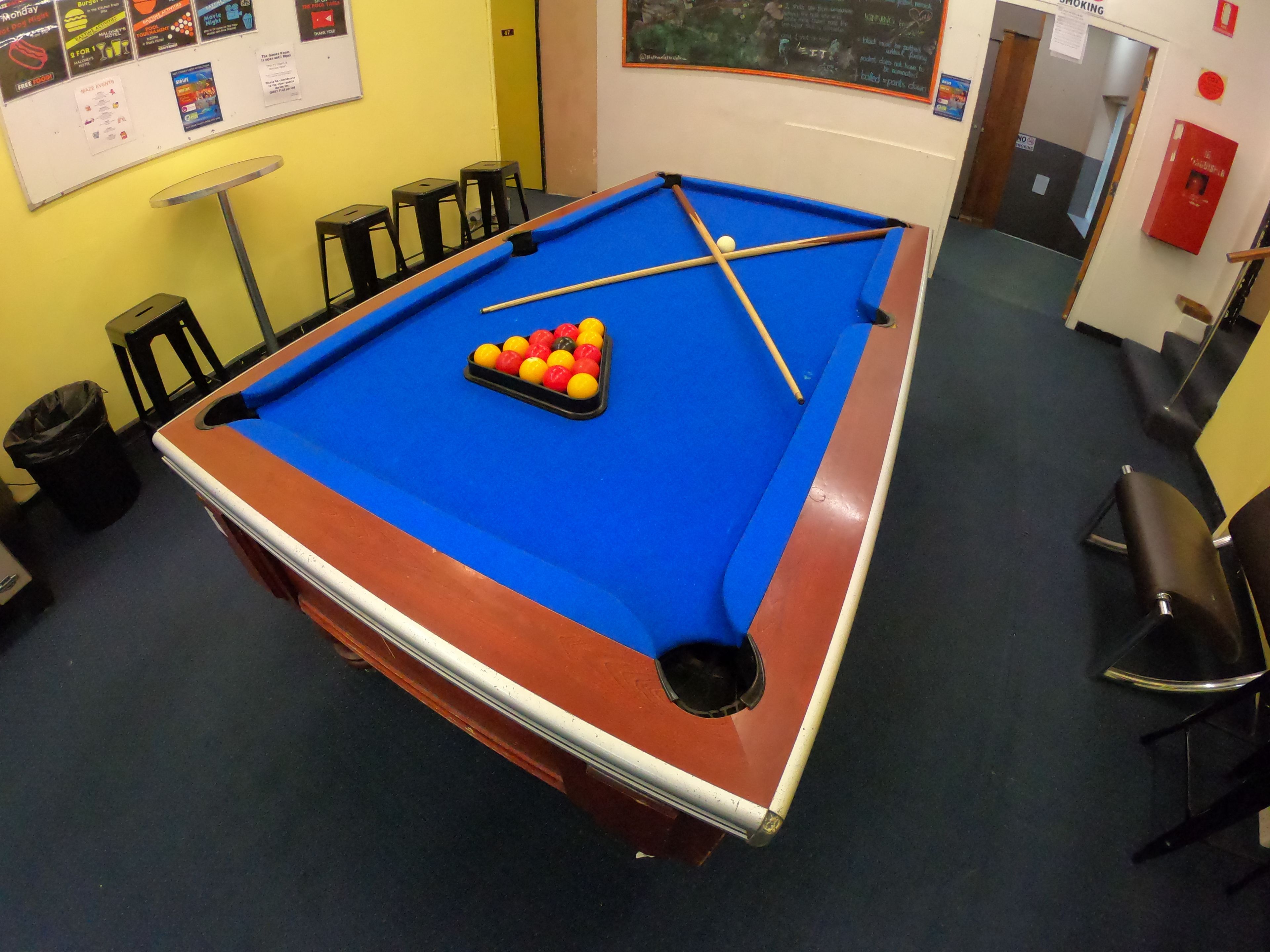 A pool table with a blue felt surface, surrounded by black stools and a yellow wall with various notices.