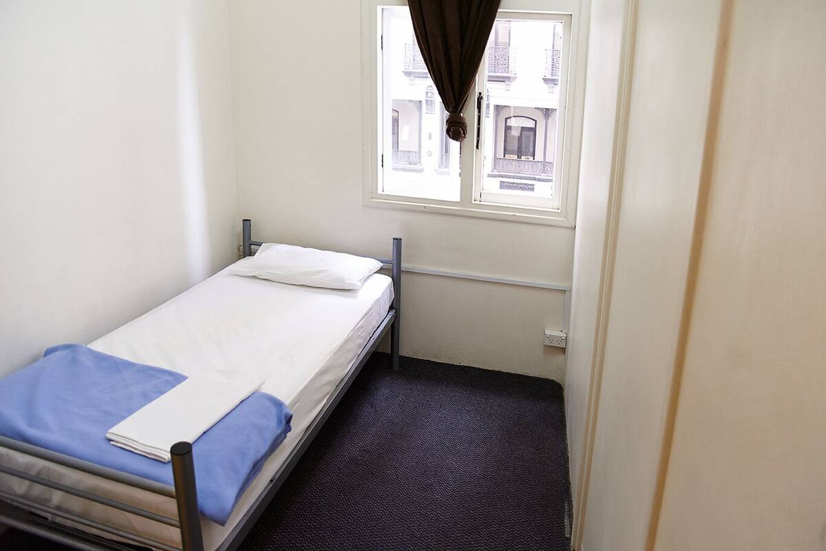 A small, single-bed room with a window, a metal bed frame, a blue blanket, and a dark carpet.