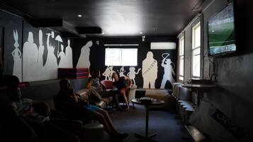 Una habitación tenuemente iluminada con una gran pantalla de televisión mostrando un partido de fútbol, una ventana con luz natural y siluetas de personas en la pared.
