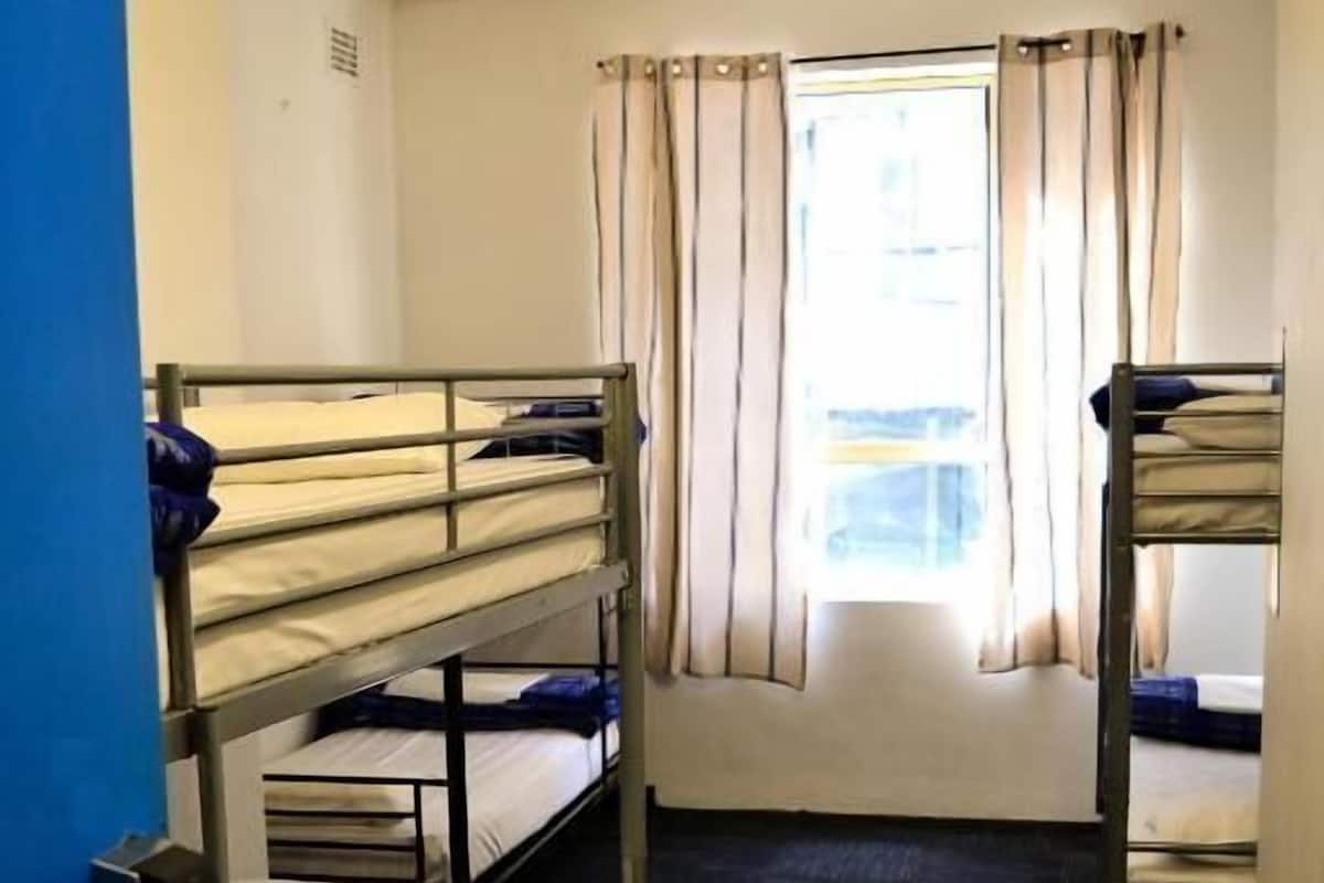 A dormitory room with bunk beds, a window with curtains, and a ceiling fan.