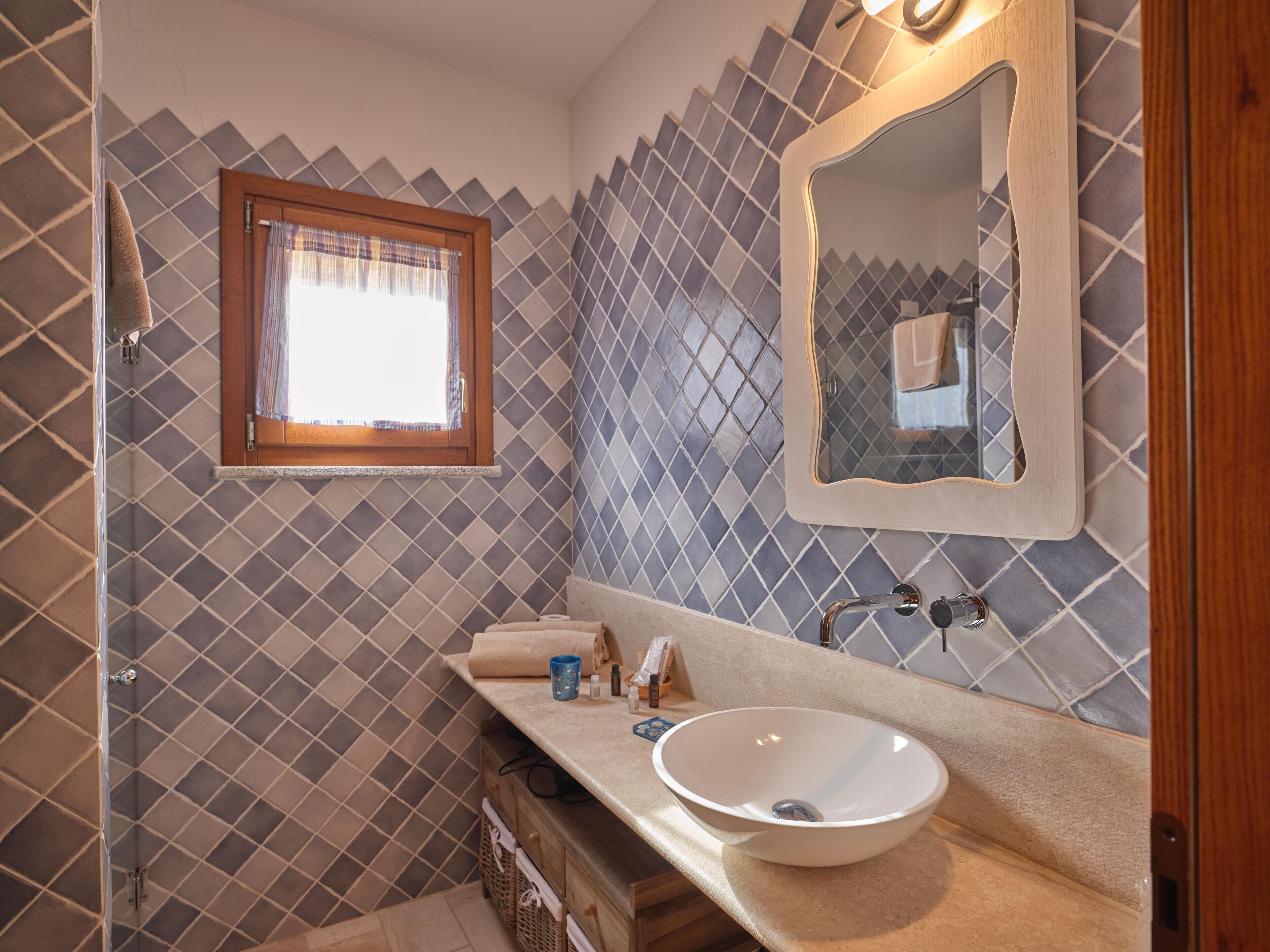 Un baño con pared de azulejos, lavamanos, espejo y ventana.
