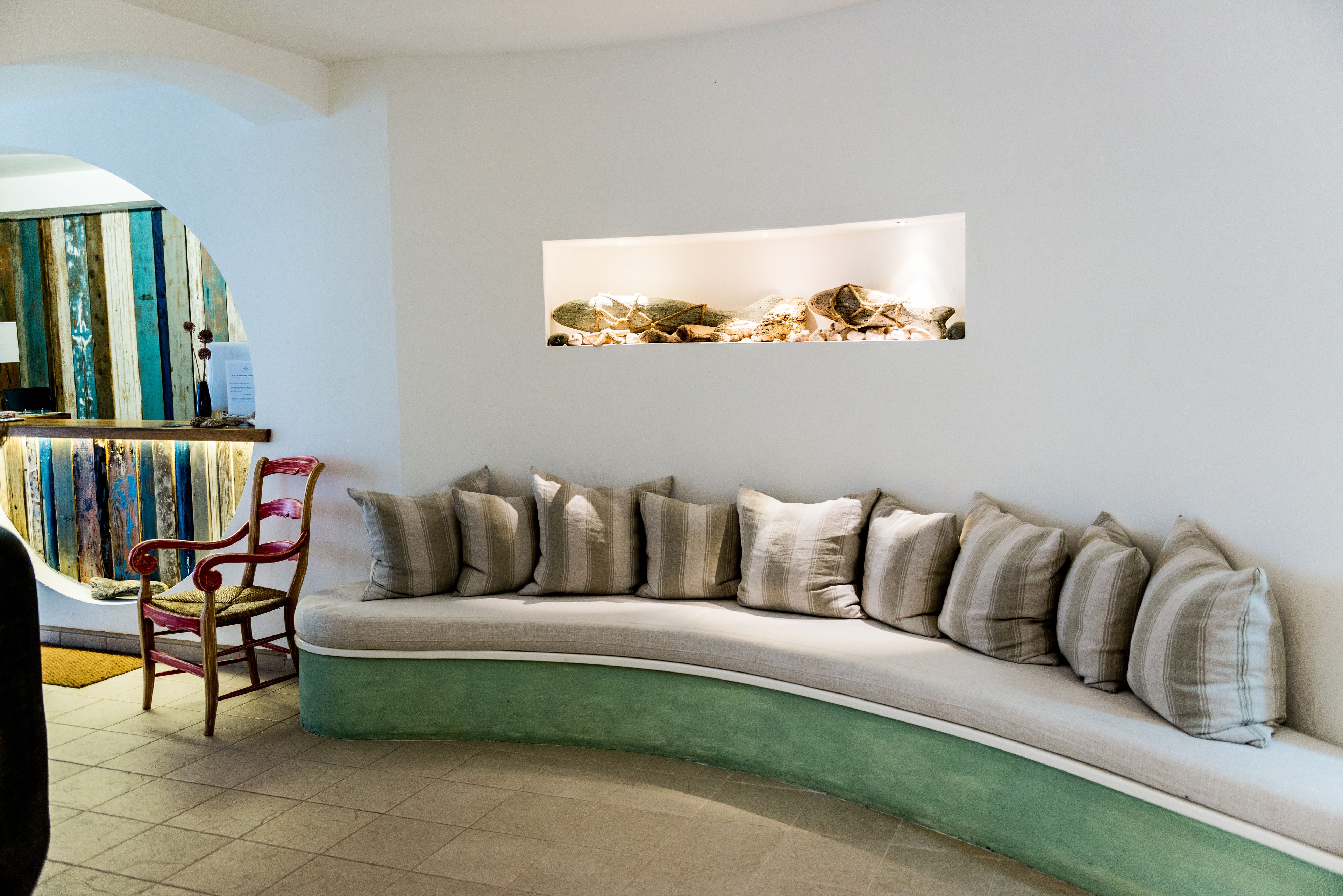 A curved sofa with multiple cushions, a wooden chair, and a wall-mounted shelf with decorative items.