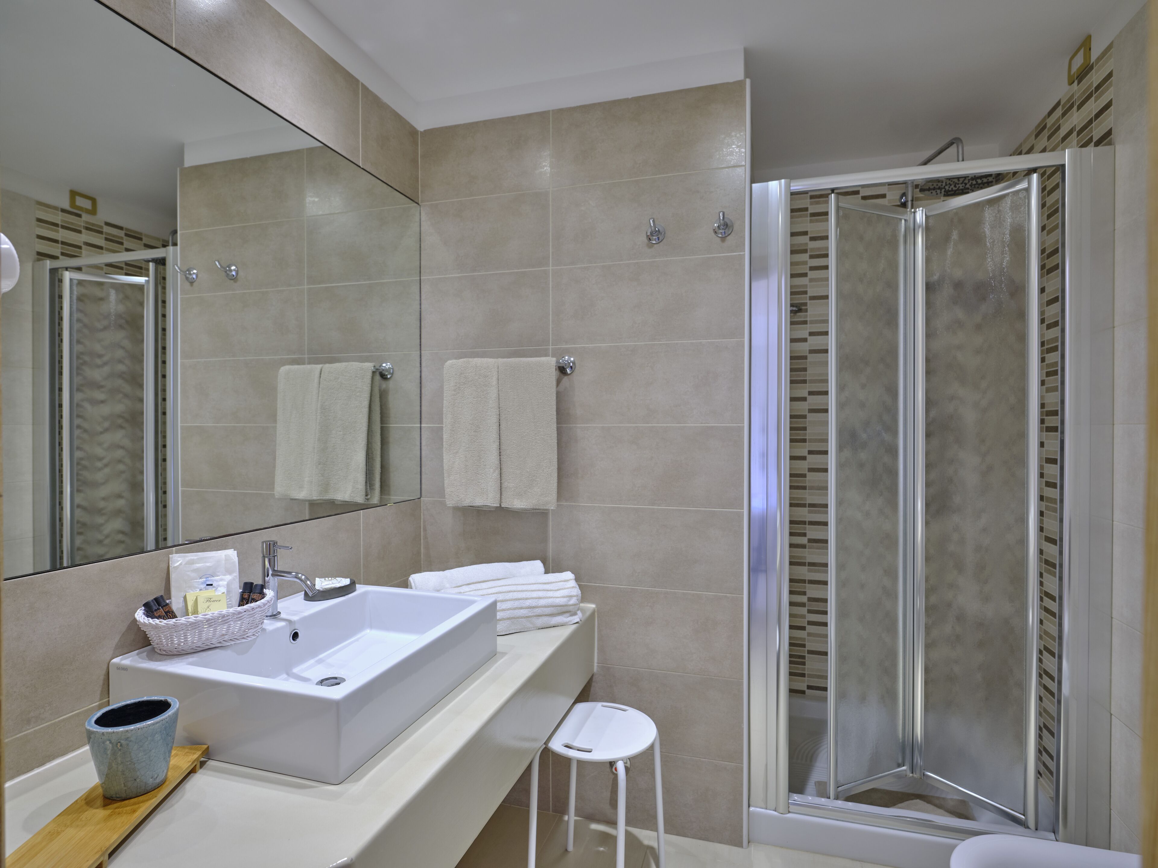 A modern bathroom with a large mirror, a sink with a basket of toiletries, two towels, and a glass shower enclosure.