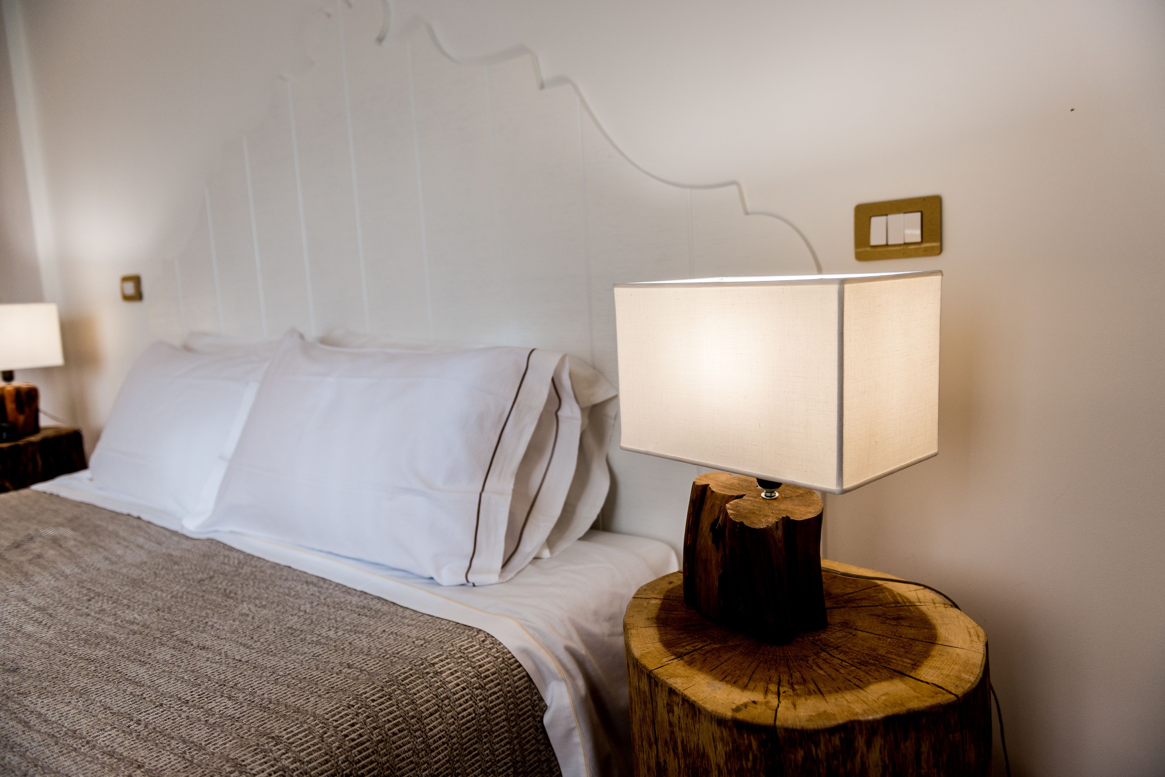 Una cama bien hecha con sábanas blancas y una manta gris, una mesita de noche con lámpara, y un interruptor de luz en la pared.