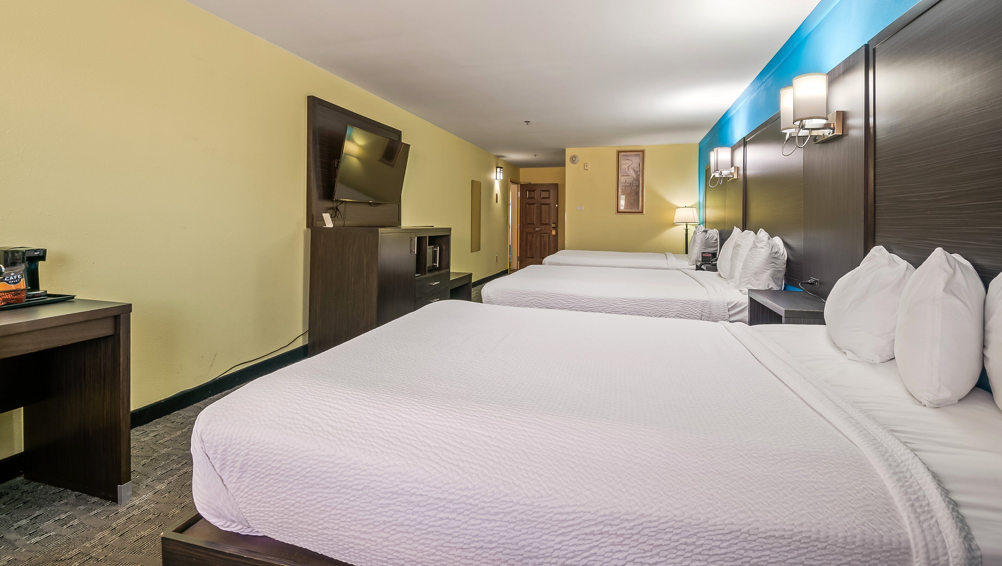 Quarto de hotel com duas camas, uma secretária com máquina de café, televisão e banheiro com espelho e iluminação.