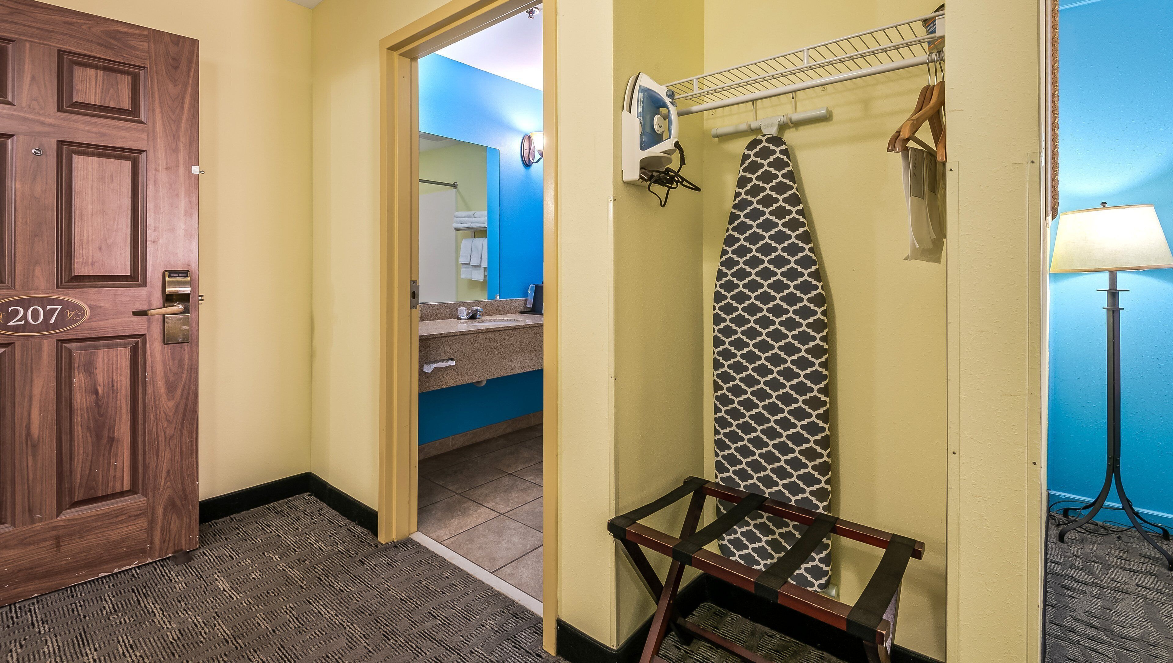A hotel hallway with a wooden door marked "207", a bathroom with a blue wall, a towel rack, and a hanging iron.