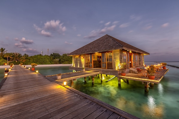 A wooden overwater bungalow with a deck, potted plants, and ambient lighting. - Komandoo Maldives Island Resort (Komandoo)