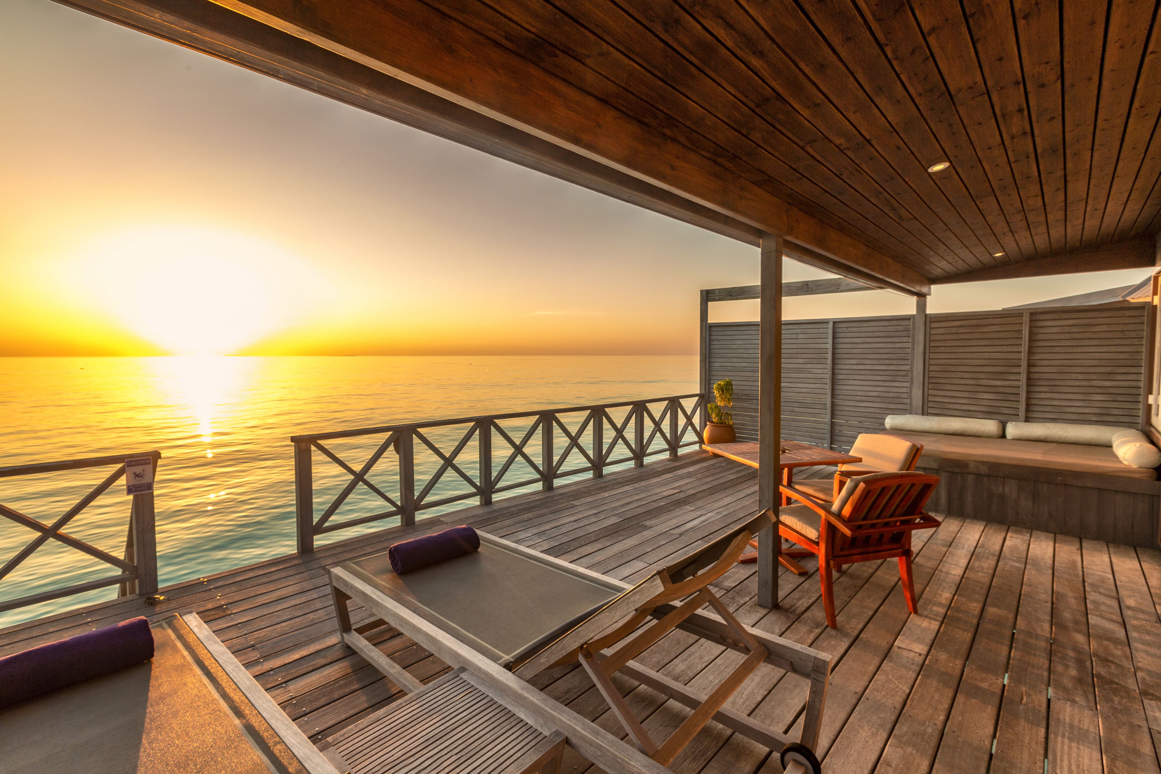 A wooden deck with a table and chairs, overlooking a sunset over the water.