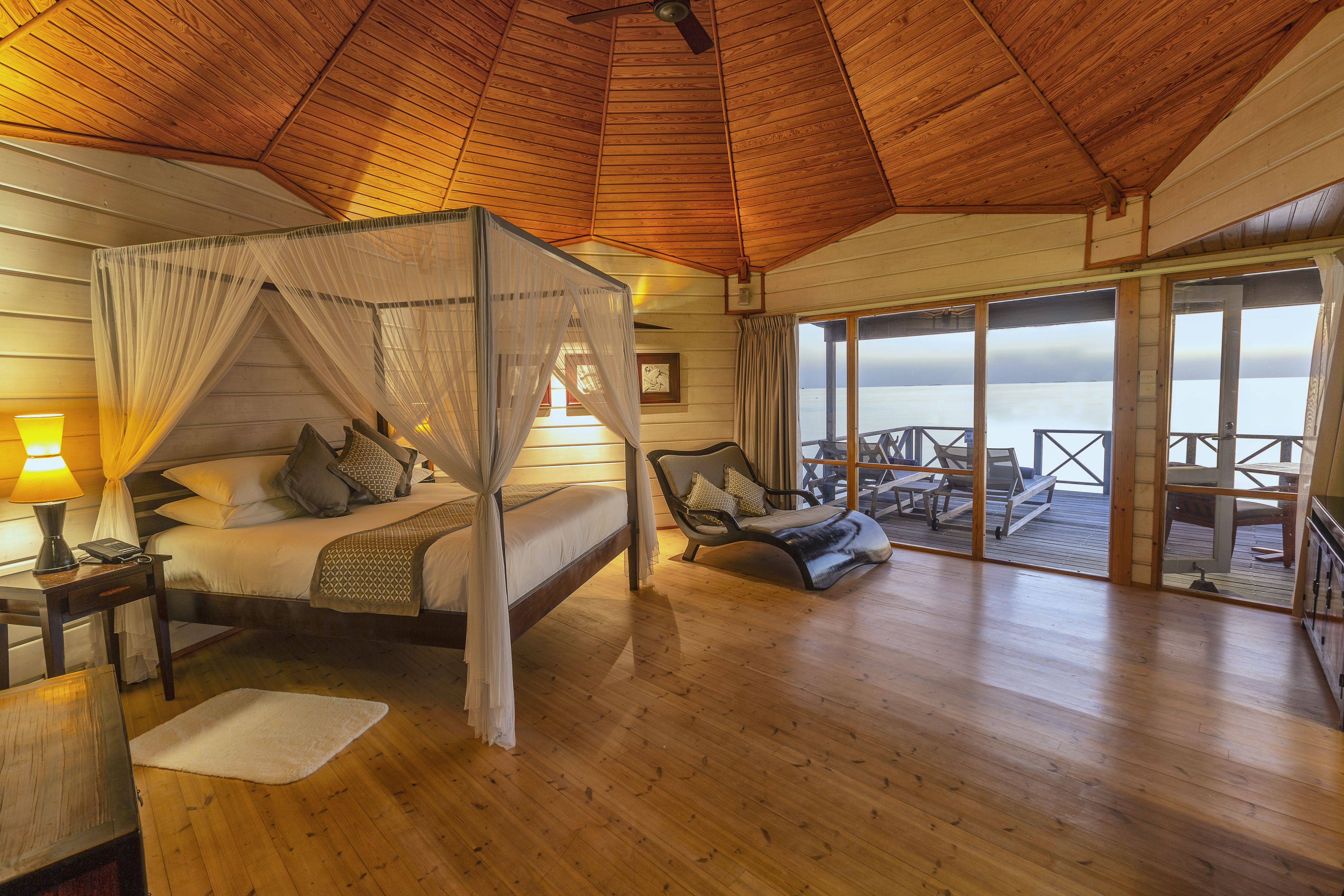 A wooden cabin with a four-poster bed, a balcony with outdoor seating, and a view of the water.