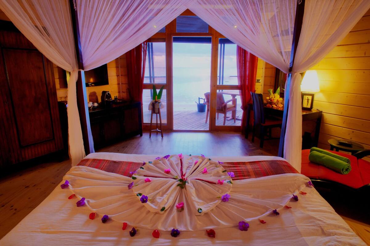 Une chambre avec un lit agrémenté d’un arrangement floral, un bureau en bois, une chaise et une vue sur la mer par une porte ouverte.