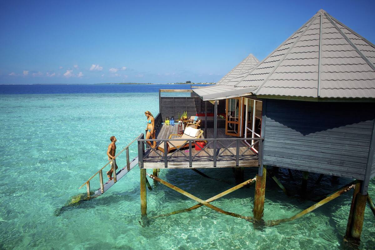 Un bungalow sur pilotis doté d’une terrasse en bois et d’un espace abrité, relié à la plage par une passerelle en bois.
