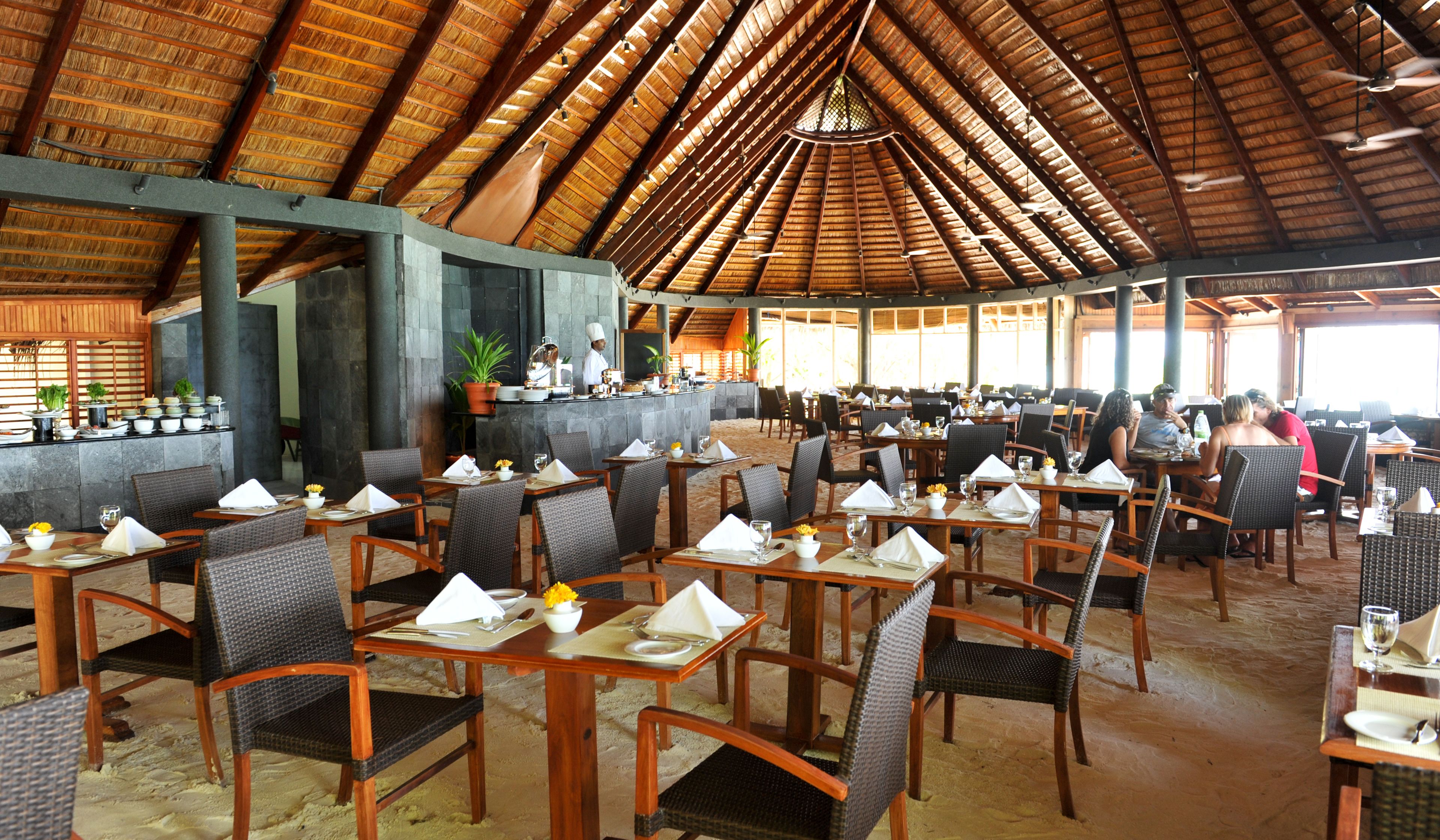 A spacious restaurant with a high thatched roof, multiple dining tables, and chairs.