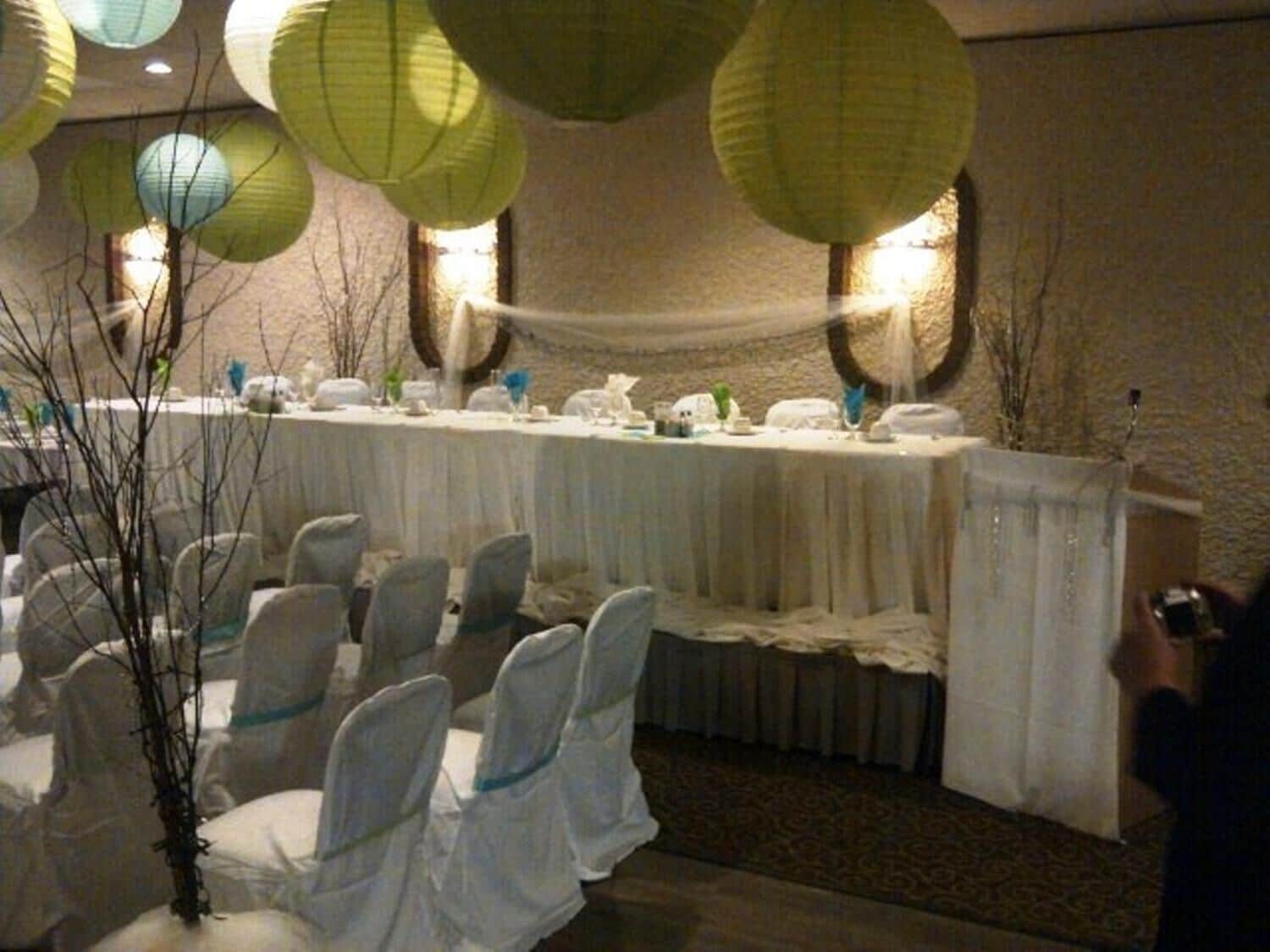 Een feestzaal met ronde tafels, bedekt met witte tafelkleden, stoelen met witte hoesjes, en lantaarns die aan het plafond hangen.