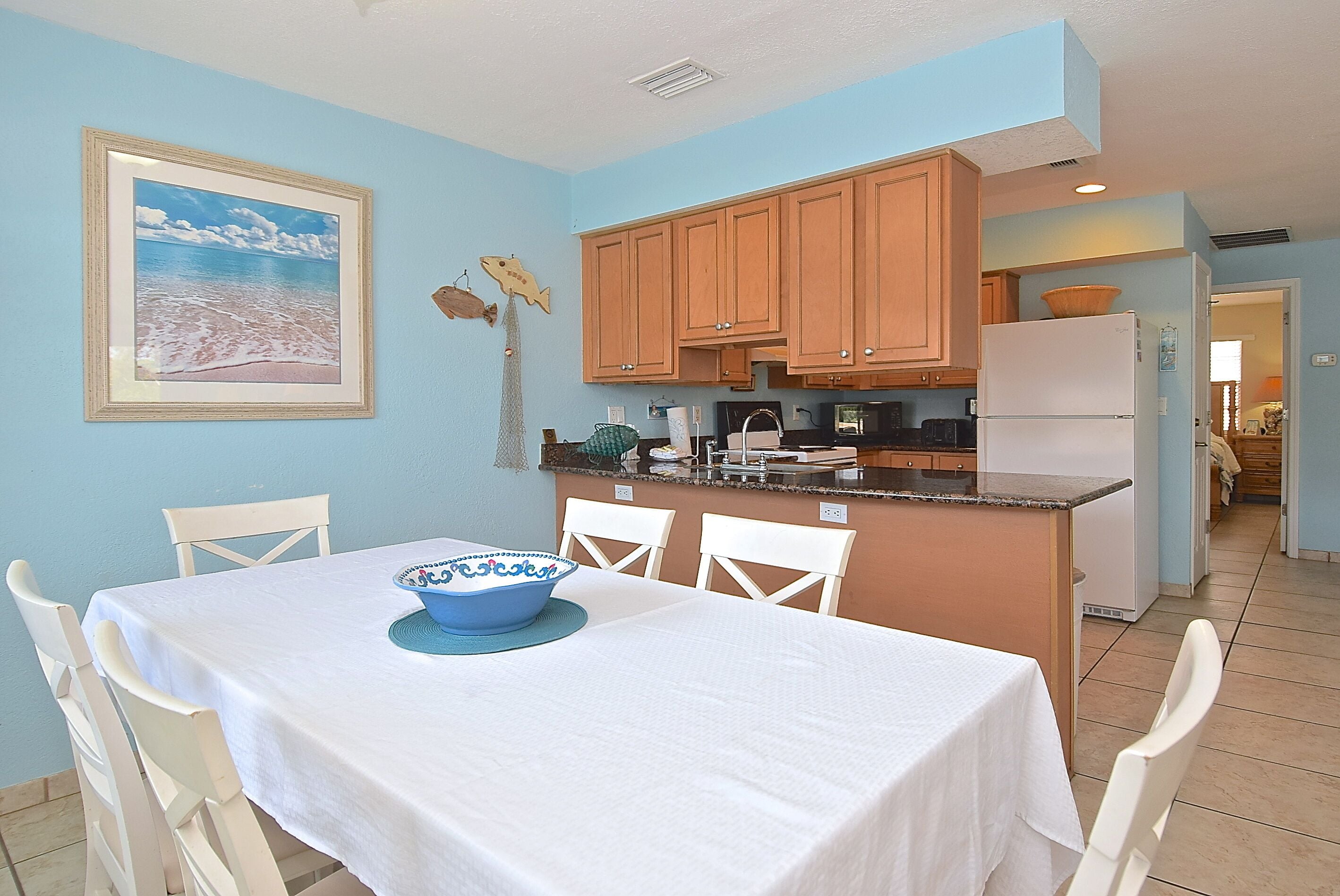 A dining area with a table set for two, a kitchen with wooden cabinets, a refrigerator, and a framed picture on the wall.