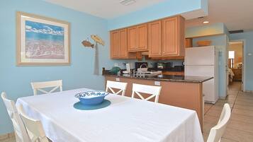 A dining area with a table set for two, a kitchen with wooden cabinets, a refrigerator, and a framed picture on the wall.